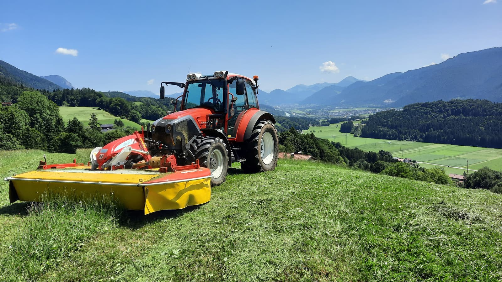 Traktor mit Mähwerk auf der steilen Bergwiese, dahinter das Inntal