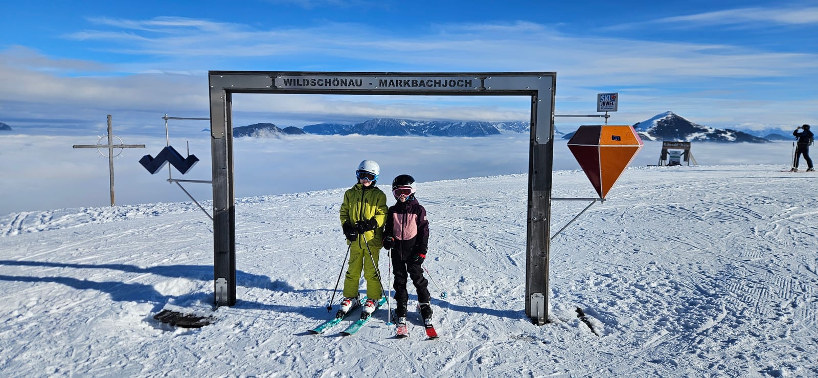 Zwei Kinder auf Skiern am Gipfeltor Wildschönau – Markbachjoch, dahinter Nebelmeer und Berge